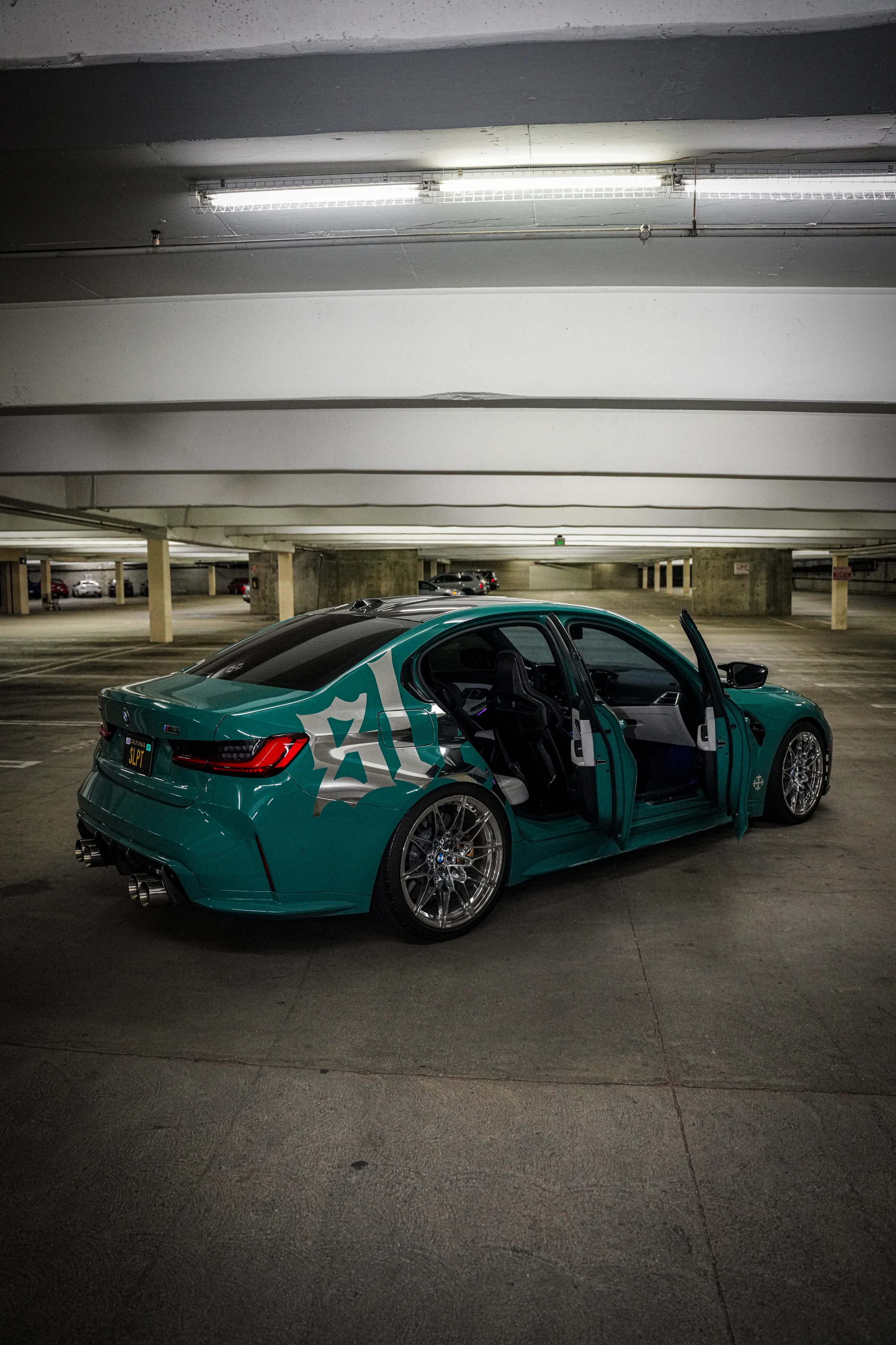 Teal SLEPT ON BMW M3 with silver graphics and polished wheels in a parking garage